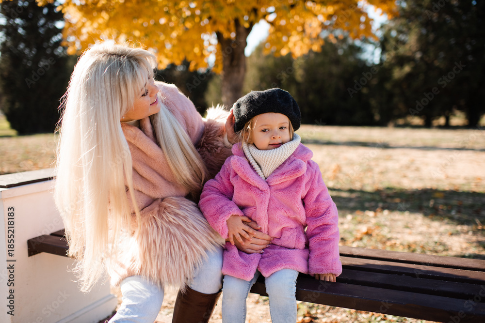 Fototapeta premium Smiling young adult woman holding child girl wearing fluffy coats over yellow leaves outdoors. Motherhood.