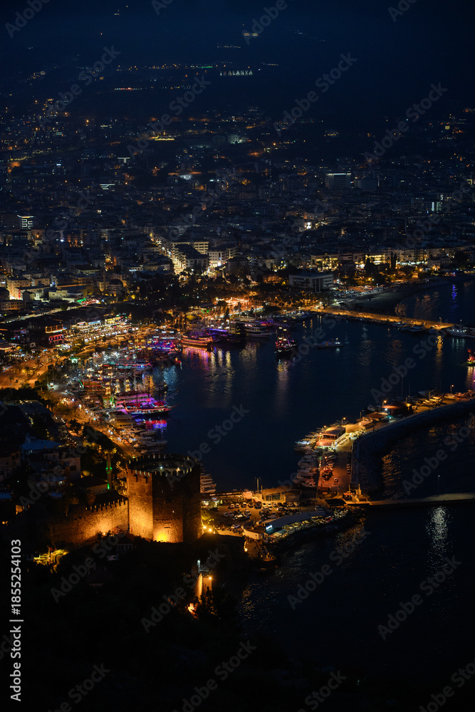 Fototapeta premium Night view of Alanya, Turkiye. Panoramic view of beach, city with street lamps, fortress walls, Red Tower, and Mediterranean Sea. Tourism and seaside recreation