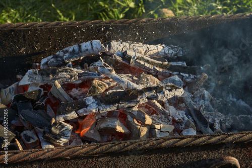 Smoldering logs and bright flames in a metal barbecue grill. Detailed close-up of fire, heat, and smoke outdoors. Flaming logs with heat and smoke rising from an open grill.