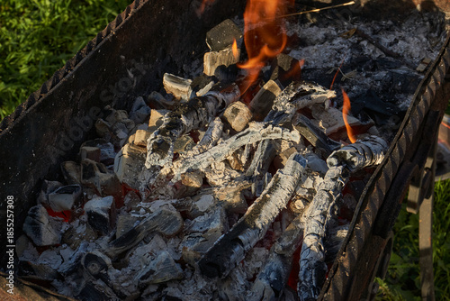 Smoldering logs and bright flames in a metal barbecue grill. Detailed close-up of fire, heat, and smoke outdoors. Flaming logs with heat and smoke rising from an open grill.