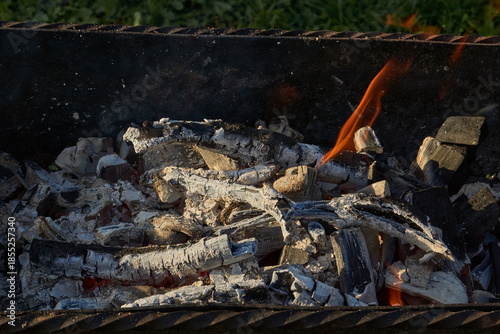 Smoldering logs and bright flames in a metal barbecue grill. Detailed close-up of fire, heat, and smoke outdoors. Flaming logs with heat and smoke rising from an open grill.