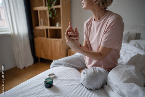 Cuadro en lienzo Middle aged woman doing beauty self care routine rituals in morning, sitting in bed and applying moisturizer lotion cream from jar on hands