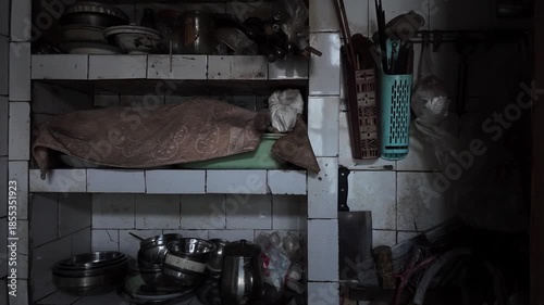 An old, cluttered kitchen storage area with open tiled shelves holding dusty cooking utensils. Household items convey a sense of aging, third-world domestic space, disorganized, poor living conditions