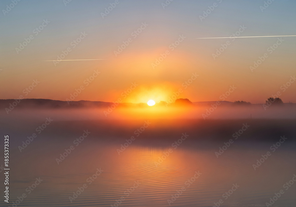 Naklejka premium Golden sunrise through morning mist over serene lake, reflecting warm light on calm waters, with distant forest, capturing environmental beauty and tranquil dawn