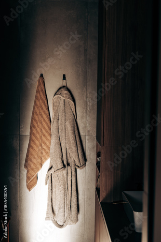 Minimalist bathroom interior with hanging bathrobe and towel, warm moody lighting and neutral textures. Cozy spa atmosphere, modern home wellness concept, winter comfort, slow living and self care.