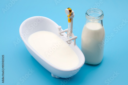 Milk Bath Skincare Concept with Mini Bathtub Filled With Milk and Glass Bottle on Blue Background
