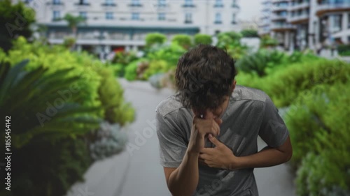Teenage boy clutching his hand to his chest and coughing on a building walkway lined with plants; distress emergency.