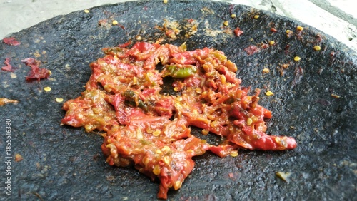 Close-up of homemade Sambal Ulek (Indonesian chili paste) freshly ground on a traditional dark stone mortar (cobek), showing spicy red chili peppers and seeds.