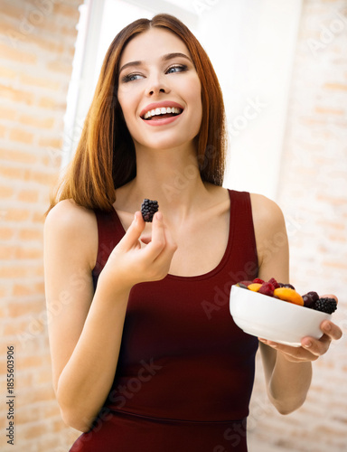 Happy smiling brunette cute woman with plate of fruits, indoors. Beautiful girl wear red dress - dieting, weight loss, healthy vegetarian eating concept.