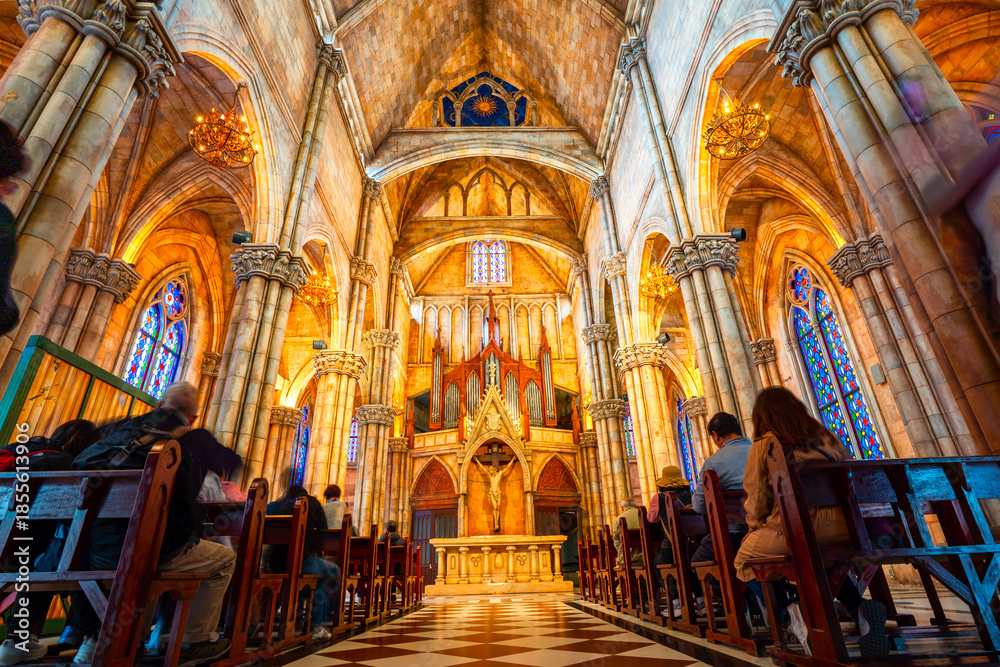Obraz premium Grand church interior featuring intricate Gothic architecture, vibrant stained glass windows, and people reverently seated in wooden pews.