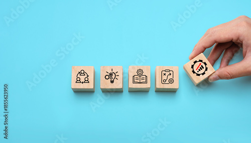 Wooden blocks with icons representing teamwork, innovation, learning, planning, and growth arranged in a row with a hand placing the final progress block, symbolizing innovation and continuous change