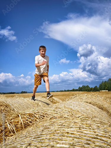 Child joyfully running over hay bales in a sunny field with blue skies and fluffy clouds in the background during a warm afternoon