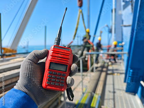 Obraz na płótnie Close-up of a red handheld radio in a worker’s gloved hand on an offshore industrial platform, with crew and crane blurred in background under clear blue sky