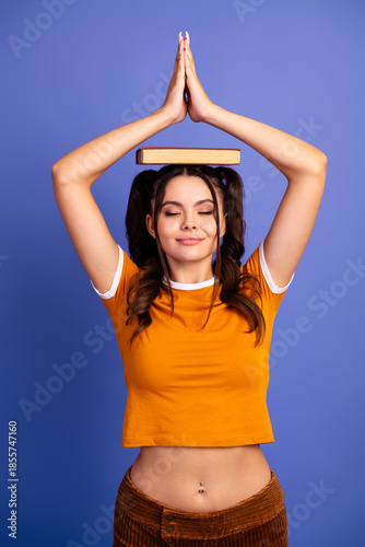 Young female teen girl balances a book on her head while practicing a calm yoga pose with hands pressed together under a violet purple background