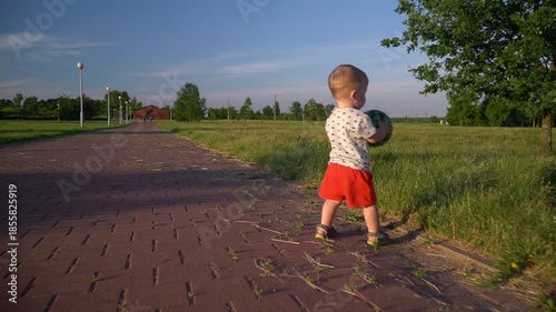 a little boy in red shorts runs along a brick path, carrying a green watermelon-patterned ball, about to throw it onto the grassy field on a sunny day.