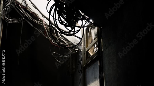 The dark concrete interior reveals exposed wiring cascading down from the ceiling, illuminated by a beam of light streaming through a window. The atmosphere is industrial and raw.
