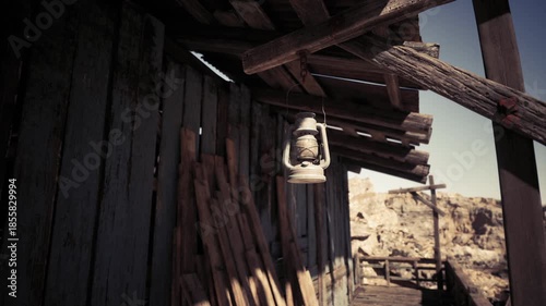 An old lantern dangles from a rustic cabin, surrounded by wooden beams and a desolate desert landscape. The scene captures the charm of a bygone era in a remote location.