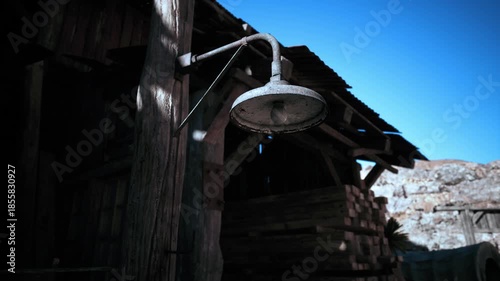 A vintage wall mounted lamp hangs beside a rustic wooden shed, casting a warm light against the weathered wood during a bright, sunny day. The scene evokes nostalgia and simplicity.