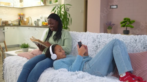Mother reading a book while her daughter is listening to music on the sofa