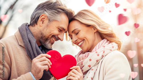 A close-up, tender portrait of a smiling middle-aged couple touching foreheads while jointly holding a small, bright red heart object, symbolizing their deep love and connection.