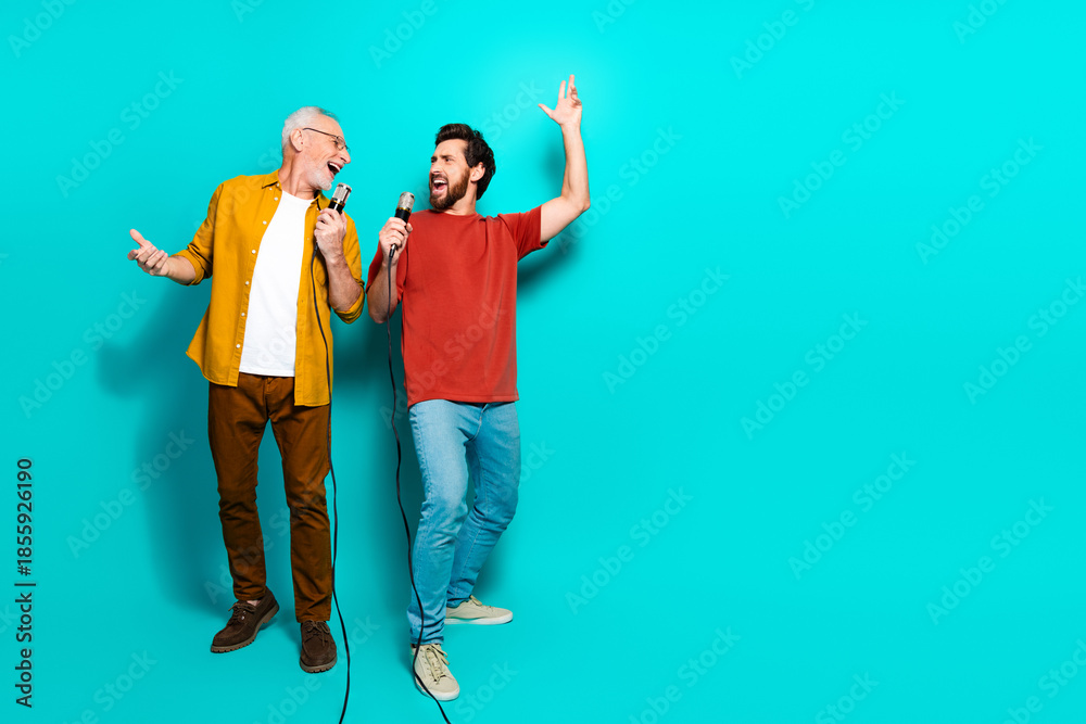 Fototapeta premium Two friends sing together in a bright studio with teal background wearing casual shirts and smiling