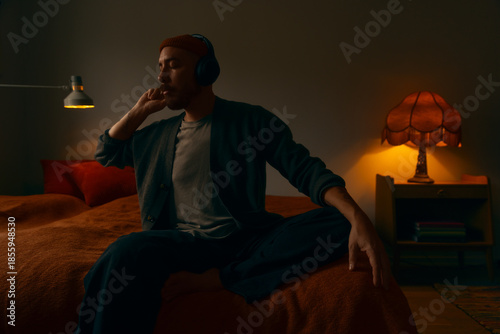 A pensive man wearing headphones and a beanie sitting on a bed in a dimly lit, cozy room. The scene features warm ambient lighting from vintage lamps, creating a moody and cinematic atmosphere