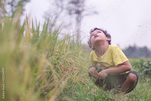 Curious young child crouching in grass and looking upward outdoors. Concept of imagination, childhood wonder, learning, mindfulness, outdoor education, and connection with nature