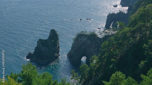 Stunning aerial footage of natural rock arches and rugged sea stacks rising from sparkling blue ocean at Kitayamazaki Cliffs in Sanriku Fukko National Park Iwate Japan.