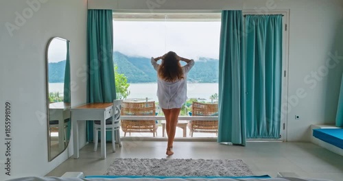 Beautiful young woman in white shirt walking bright hotel room, relax and enjoy serene morning landscape lake view from a wide window, relaxation, travel, and vacation lifestyle in Bali. Back view