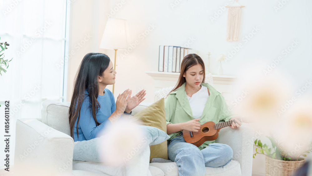 Obraz premium Two young Asian women sharing a joyful moment at home. One girl is playing the ukulele while her friend claps along and cheers. They are sitting on a cozy sofa in a bright, minimalist living room.