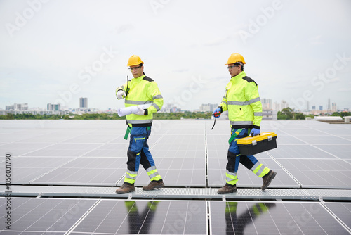 Two construction workers walking on a rooftop solar panel array, one carrying a blueprint, another with a toolbox. They wear safety gear, indicating a professional environment