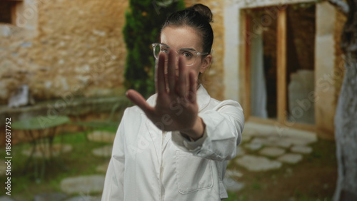 Young hispanic woman scientist in white lab coat raises palm stop gesture at stone building entrance; denial.
