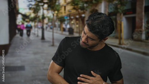 Young hispanic man grasping his stomach with both hands on a busy city street lined with buildings; discomfort.