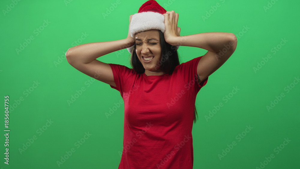 Naklejka premium Young hispanic woman wearing santa hat and red shirt with hands on head and squinting face in studio on green screen; holiday frustration.