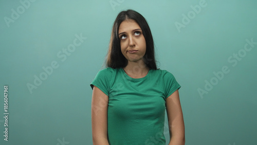 Photos Young hispanic woman in green t shirt rolling eyes and pursing lips while standing and facing camera in studio with teal backdrop; annoyance