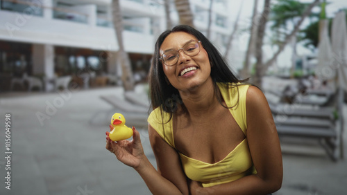Fotografi Woman holding rubber duck up while smiling, cleavage visible at resort building poolside with loungers and palm trees; playful leisure moment
