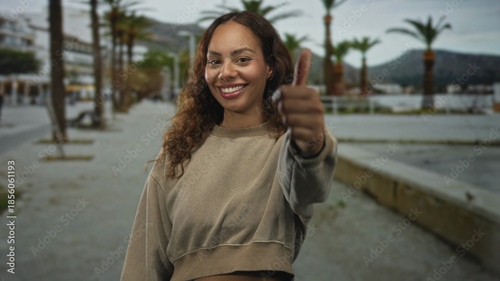Fototapeta premium Young african american woman showing thumbs down toward camera on a palm lined street by the waterfront, neutral expression; disapproval.