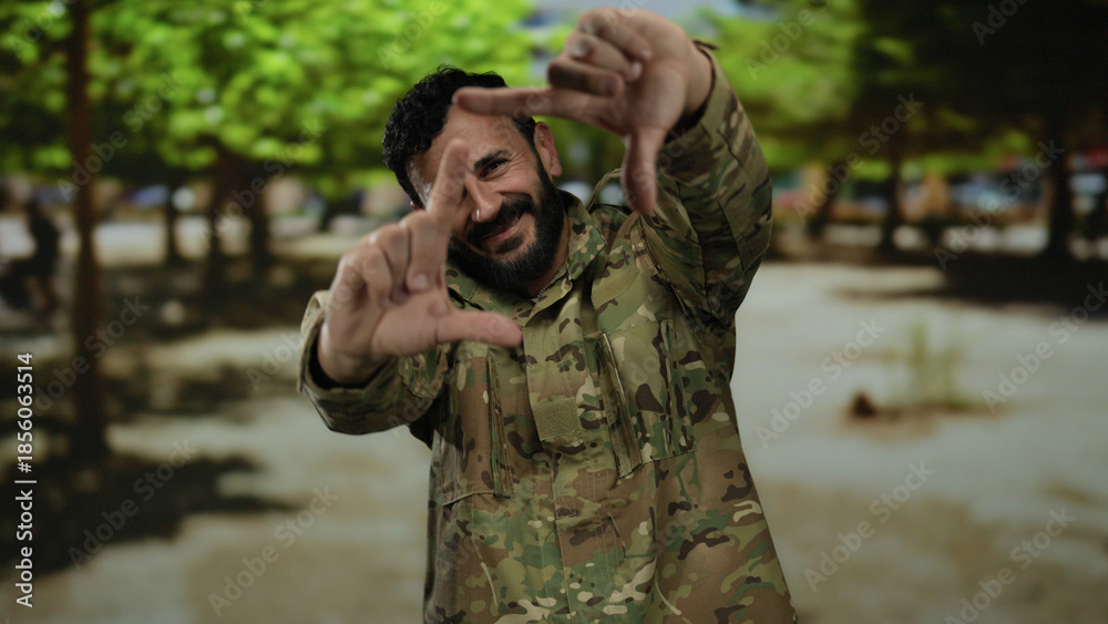 Obraz premium Hispanic man in military uniform frames his view with hands while standing on a street lined with trees, suggesting creativity outdoors.