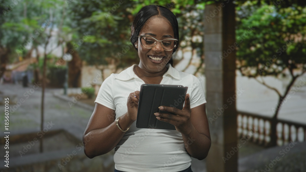 Fototapeta premium Woman holding tablet with fist raised and smiling, wearing glasses and bracelet, standing on street; confidence success.