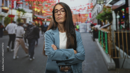 Vászonkép Young woman with glasses and crossed arms on crowded street, wearing blazer and visible belt, looking defiant; confidence resolve