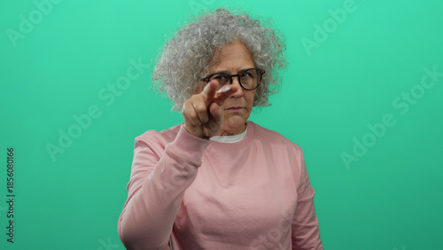Elderly woman with grey hair gesturing vigilance against a green background, wearing glasses and a pink sweater, adds an intriguing focus to social awareness.