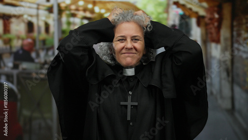 Quadro su tela Elderly woman with grey hair and clerical attire poses outdoors on a bustling restaurant terrace, showcasing serenity and faith in an urban exterior setting
