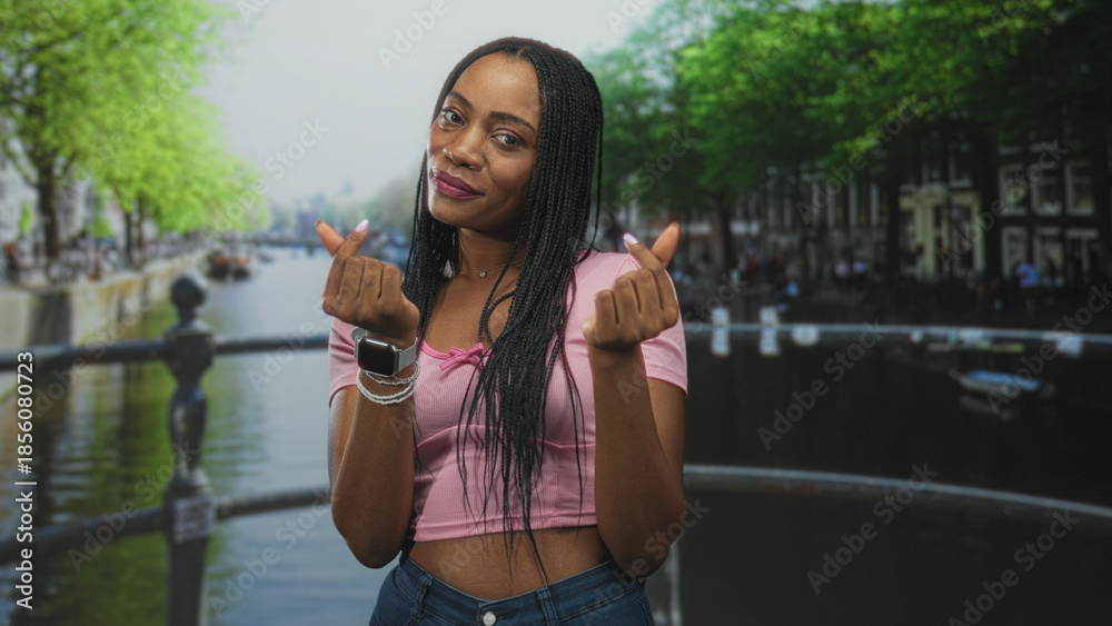 Fototapeta premium Black woman on a canal bridge making finger heart gestures with hands and visible midriff in a pink crop top; playful affection.