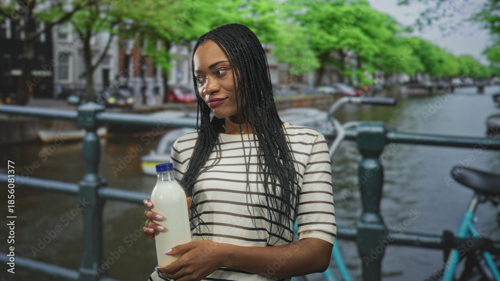 Fototapeta premium Woman holding milk bottle in hand on street by canal, striped shirt and braided hair visible; serenity sustainability.
