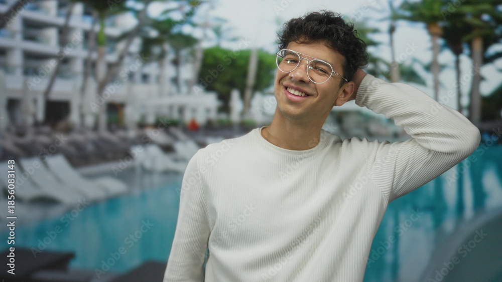 Fototapeta premium Man smiles while tilting head at resort pool adjacent to hotel building with palm trees in view; relaxation.