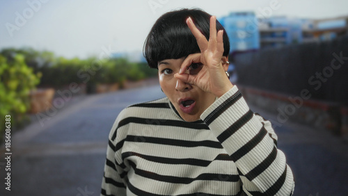 Fotografi Young chinese woman playfully making an ok gesture over her eye while standing in a city street, wearing a striped sweater, embodying urban and youthful outdoor vibes