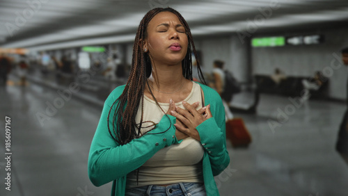 Fotografie Woman clutching chest in airport terminal, hands pressed to chest in apparent heartattack with strained face and closed eyes; panic emergency