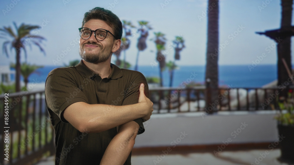 Fototapeta premium Man in brown polo shirt holds and rubs bare forearm on building balcony overlooking palm trees and ocean; discomfort seaside scene.