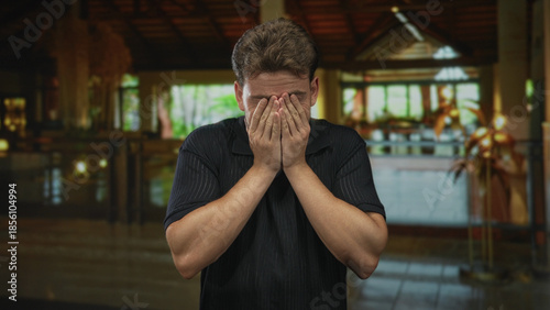 Bild auf Leinwand Man with hands covering face and head bowed in hotel building lobby wearing black polo shirt; frustration reflection