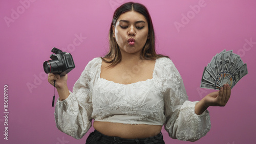 Fényképezés Woman holding a dslr camera and fanning a stack of us dollar bills with puckered lips in a pink studio; confidence monetization business